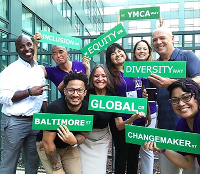 Eight members of YMCA of the USA's Global Diversity, Equity & Inclusion team holding signs that say "YMCA," "Baltimore," "equity," "inclusion," "diversity," "global," and "changemaker."